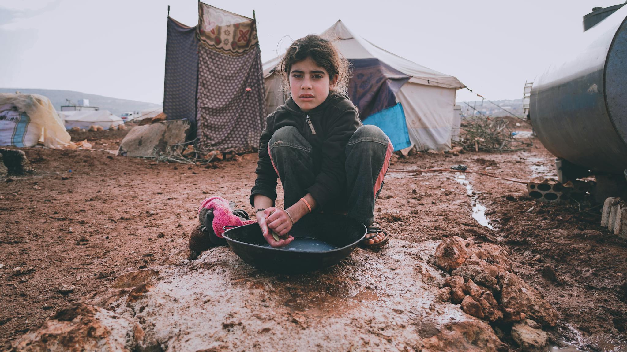 A young girl cleans dishes in a refugee camp, highlighting resilience.